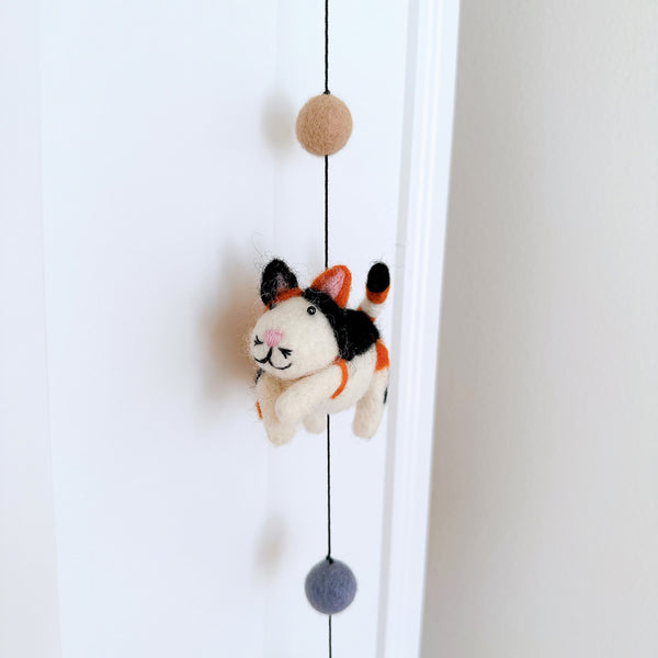 A charming 3D needle-felted cat from a Felt Cat Garland hangs against a bright white door. Crafted from 100% wool, the white felt cat features distinct black, orange, and white markings, small pink ears, a stitched pink mouth, and matching pink paws. It is suspended by a black string with a tan felt ball above and a gray one below, emphasizing its whimsical, handcrafted design.