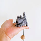 A striped wool felt cat, a charming part of a Felt Cat Garland, rests in an open palm against a stark white background. This playful cat features defined stripes, small upright ears, and a petite tail. A complementary tan, spherical felt bell dangles by a string below the handcrafted cat.