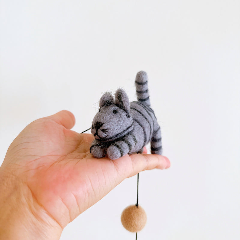 A striped wool felt cat, a charming part of a Felt Cat Garland, rests in an open palm against a stark white background. This playful cat features defined stripes, small upright ears, and a petite tail. A complementary tan, spherical felt bell dangles by a string below the handcrafted cat.