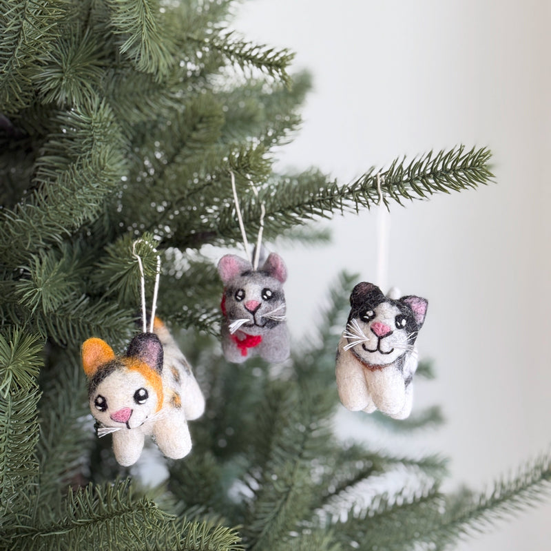 Three charming handmade felt cat ornaments – a Calico, a Gray Tabby, and a Tuxedo – adorn green Christmas tree branches. Each unique wool felt ornament is meticulously handcrafted, featuring distinct details like the Calico's orange, black, and white patches, the Gray Tabby's gray fur and red scarf, and the Tuxedo's classic black and white design. All showcase sweet white whiskers, pink noses, and small black eyes.