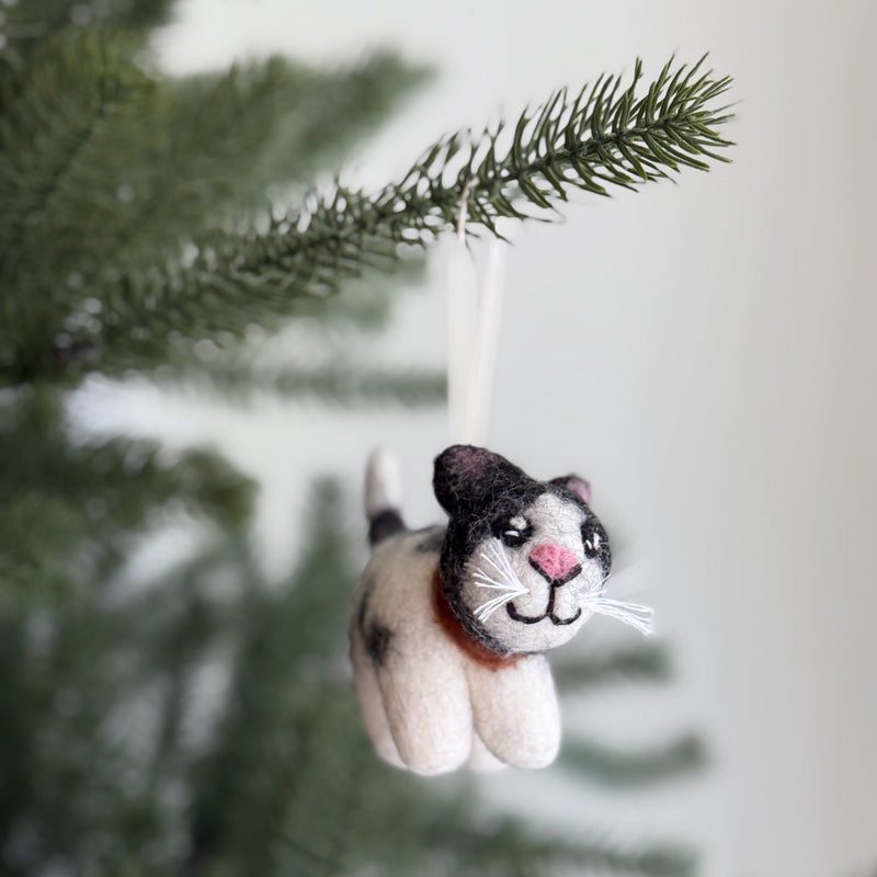 A Calico felt cat ornament hangs on a Christmas tree branch against a soft white background. This handcrafted felt cat is made from 100% New Zealand wool, featuring a white body with gray and black markings, a pink nose, a stitched smile, and white whiskers. A white string is attached for hanging, adding a whimsical touch.
