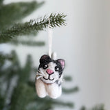 A handcrafted wool felt calico cat ornament hangs delicately from a pine branch against a soft, neutral backdrop. This felted cat features distinct black and white markings, bright eyes, a tiny pink nose, and white whiskers, all crafted from soft, textured felt.