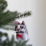 A wool felt gray tabby cat Christmas ornament hangs on a lush green tree branch, setting a cozy, festive scene. This handcrafted felt cat ornament displays a charming mix of gray, white, and black, accented by a bright red collar. Expressive black eyes and delicate white whiskers add whimsy, while a white cotton string allows it to dangle gracefully.