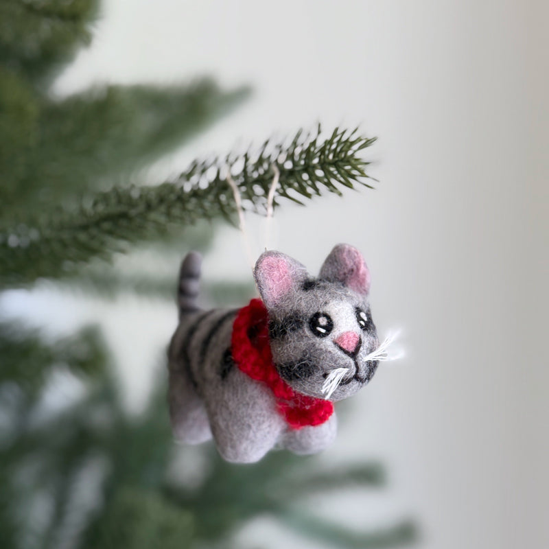 A handcrafted wool felt gray tabby cat ornament is suspended from a fir tree branch, creating a festive display. This unique Christmas ornament, made from 100% New Zealand wool, showcases a gray tabby cat with distinct gray and darker gray stripes on its body and tail. A cheerful red scarf completes the cat's look.