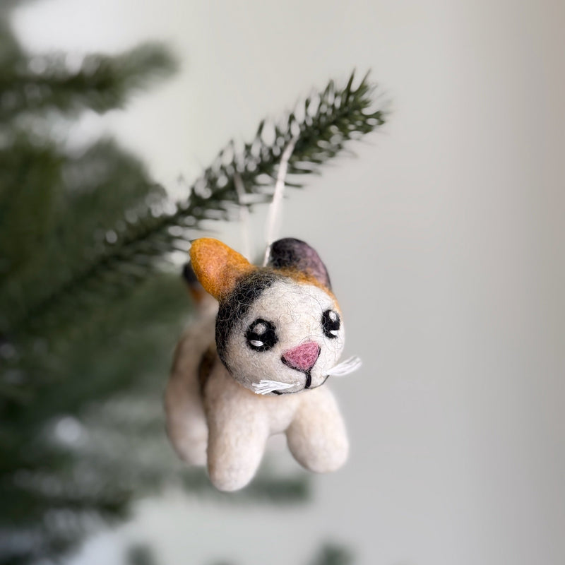 A charming Calico felt cat ornament, with its cream body patched in brown and black, hangs from a white string against a blurred background of green pine needles and a light gray wall. This wool felt cat ornament features a pink triangular nose and delicate white whiskers, adding a lifelike charm. It's ready to adorn any Christmas tree.