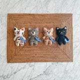 Four wool felt cat ornaments, each a stitched cat holding a blue felt fish, rest in a row on a rectangular, woven placemat, set against a white marble background. The adorable feline ornaments showcase distinct color variations: white with orange and black stripes, grey with darker grey stripes, tan and brown stripes, and solid black. Each charming handmade felt cat ornament features pointed ears and intricately stitched facial details, creating a whimsical and decorative appeal.