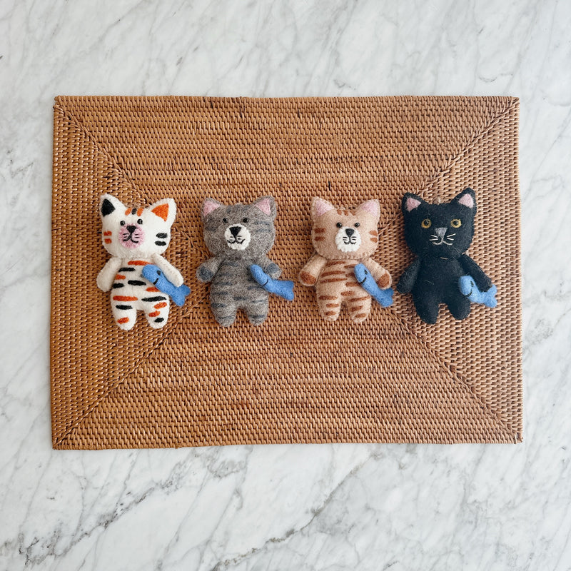 Four wool felt cat ornaments, each a stitched cat holding a blue felt fish, rest in a row on a rectangular, woven placemat, set against a white marble background. The adorable feline ornaments showcase distinct color variations: white with orange and black stripes, grey with darker grey stripes, tan and brown stripes, and solid black. Each charming handmade felt cat ornament features pointed ears and intricately stitched facial details, creating a whimsical and decorative appeal.