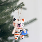 A handcrafted felt cat ornament, designed as a white cat with alternating black and orange stripes, hangs playfully from a pine tree branch. This charming stitched cat ornament, with black eyes and a pink nose, holds a blue felt fish in its arms. A jute string attached for hanging completes this festive and unique Christmas ornament.