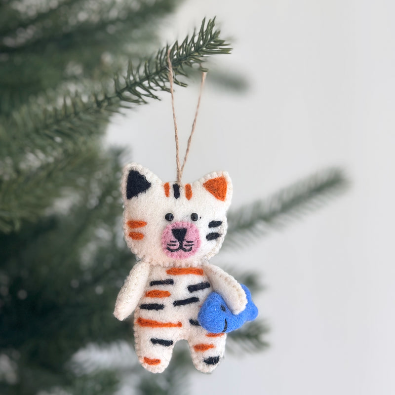 A handcrafted felt cat ornament, designed as a white cat with alternating black and orange stripes, hangs playfully from a pine tree branch. This charming stitched cat ornament, with black eyes and a pink nose, holds a blue felt fish in its arms. A jute string attached for hanging completes this festive and unique Christmas ornament.