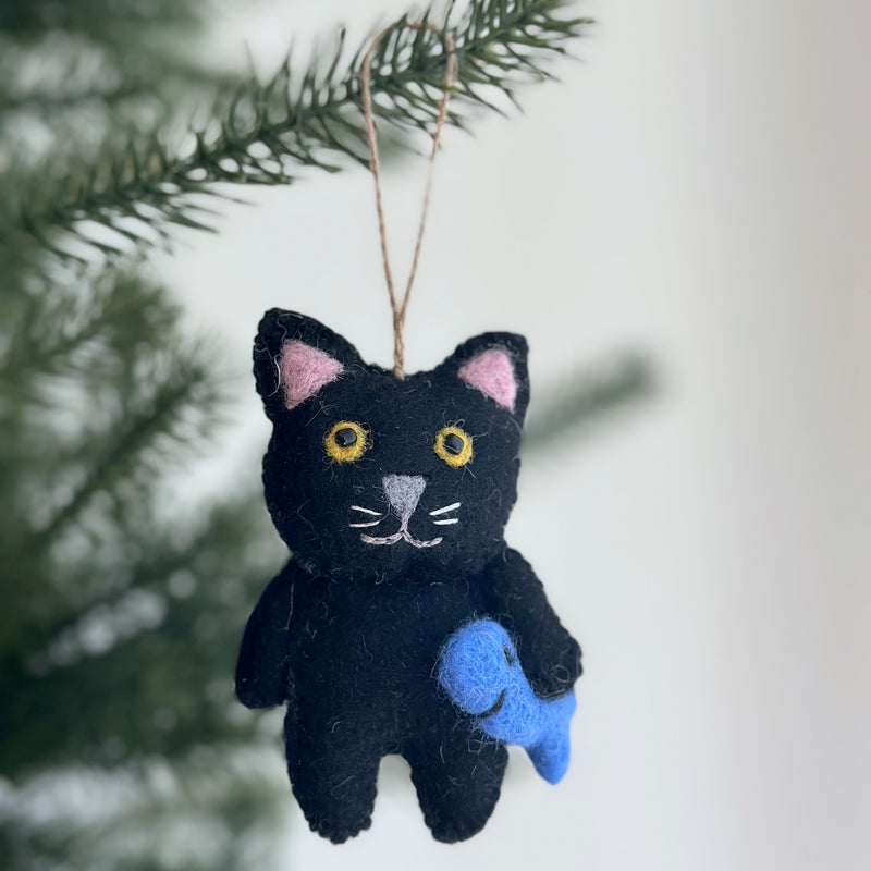 A hand-stitched black felt cat ornament, holding a blue felt fish, hangs on a softly lit Christmas tree. This charming wool felt cat ornament features pink inner ears, yellow eyes, a gray nose, and embroidered white whiskers. A jute string is attached for hanging.
