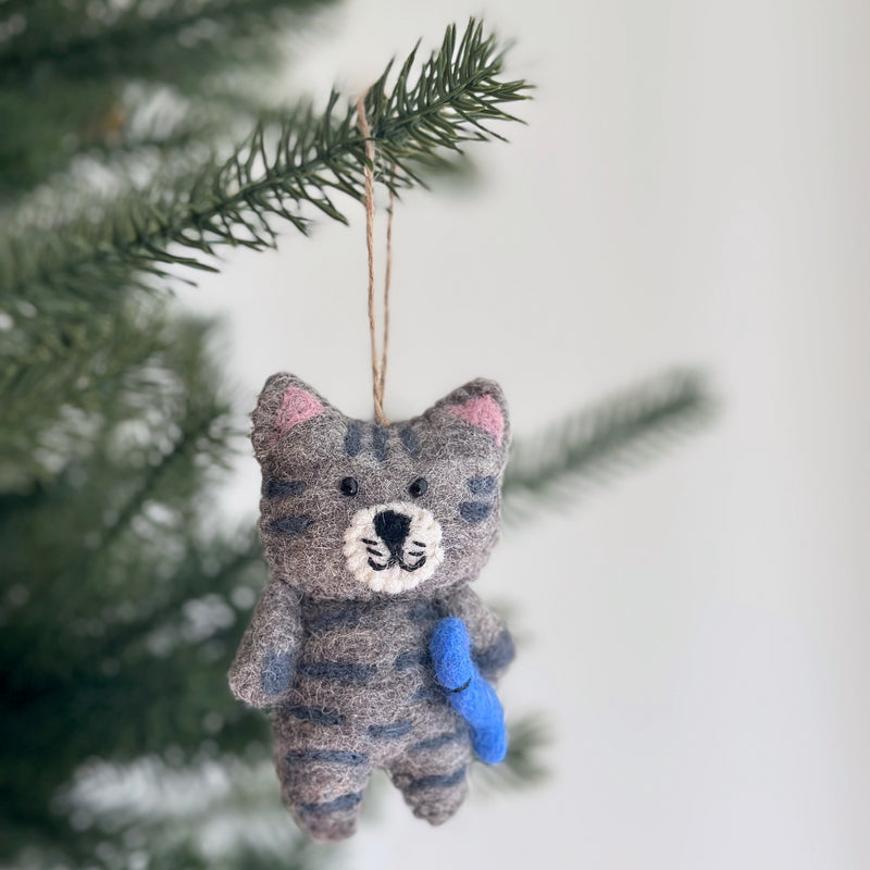 A charming wool felt cat Christmas ornament, handcrafted with a stitched tabby-like gray body and darker stripes, dangles from a pine tree branch. This festive feline features small pink inner ears, a white stitched mouth, and lovingly clutches a bright blue felted fish.