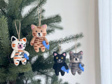 A handcrafted Wool Felt Stitched Cat Christmas ornament, featuring intricate hand-stitched details, hangs from a jute string on a Christmas tree. This unique felt cat ornament depicts a cat holding a blue fish, with delicate embroidery highlighting its feline characteristics. It adds a cozy touch to the festive setting.