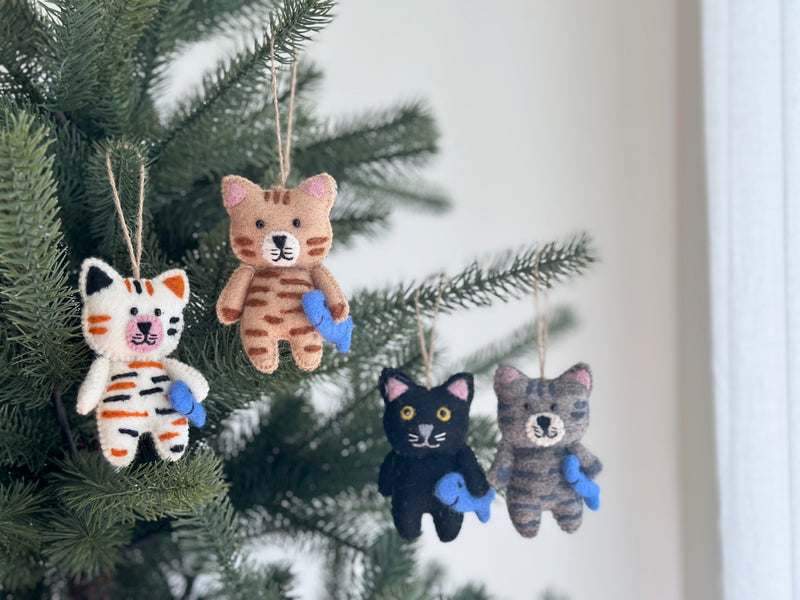 A handcrafted Wool Felt Stitched Cat Christmas ornament, featuring intricate hand-stitched details, hangs from a jute string on a Christmas tree. This unique felt cat ornament depicts a cat holding a blue fish, with delicate embroidery highlighting its feline characteristics. It adds a cozy touch to the festive setting.