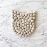 A light gray felt cat coaster rests on a light marble surface, creating a calm and minimalist atmosphere. This handmade felt coaster features subtle cat ears and is crafted from eco-friendly wool felt balls, hand-stitched for durability and a cozy, whimsical touch. It is approximately 4 inches in diameter and 0.5 inches thick.