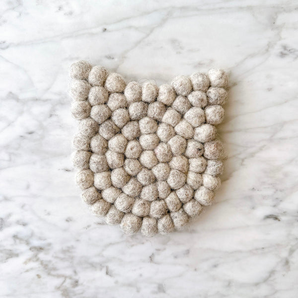 A light gray felt cat coaster rests on a light marble surface, creating a calm and minimalist atmosphere. This handmade felt coaster features subtle cat ears and is crafted from eco-friendly wool felt balls, hand-stitched for durability and a cozy, whimsical touch. It is approximately 4 inches in diameter and 0.5 inches thick.