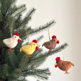 Four felt chicken ornaments, in white, yellow, brown, and beige, are displayed hanging on a vibrant green Christmas tree. Each handmade felt chicken ornament features a red comb, yellow beak, and orange feet. Suspended by rustic jute strings, these unique Christmas ornaments create a joyful and whimsical effect.