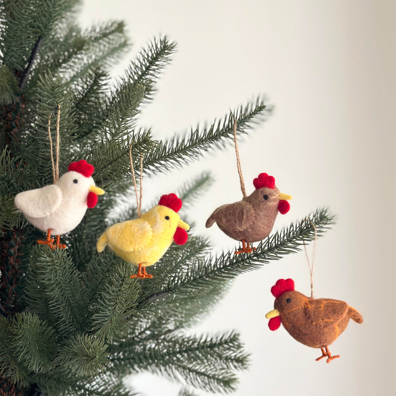 Four felt chicken ornaments, in white, yellow, brown, and beige, are displayed hanging on a vibrant green Christmas tree. Each handmade felt chicken ornament features a red comb, yellow beak, and orange feet. Suspended by rustic jute strings, these unique Christmas ornaments create a joyful and whimsical effect.