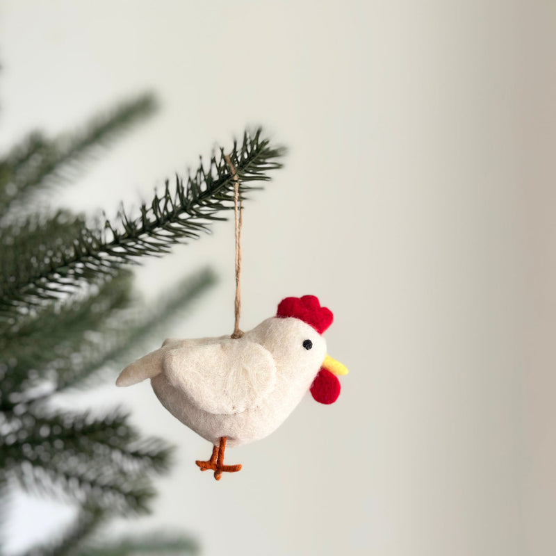 A handcrafted felt chicken ornament is gracefully displayed on a Christmas tree branch, set against a soft white backdrop. This charming white felt chicken features a vibrant red comb, a bright yellow beak, and distinct orange legs. A jute string is attached for easy hanging, highlighting its unique artisanal quality.