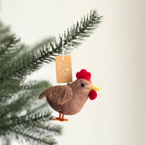A brown felt chicken ornament hangs from a pine branch, creating a warm, festive scene. This handmade felt chicken ornament is shaped like a chicken, featuring a bright red comb and wattle, and orange feet. A small tag is tied to its cotton string hanger.