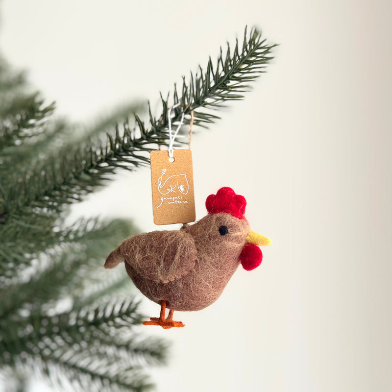 A brown felt chicken ornament hangs from a pine branch, creating a warm, festive scene. This handmade felt chicken ornament is shaped like a chicken, featuring a bright red comb and wattle, and orange feet. A small tag is tied to its cotton string hanger.