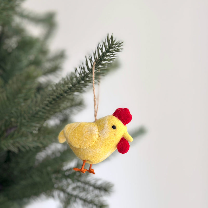 A vibrant yellow felt chicken ornament hangs from a branch of a green Christmas tree, positioned against a light, clean background. This cheerful handcrafted felt chicken features a bright yellow body, accented by a red comb and wattle, and orange feet. A jute string is attached for easy hanging.