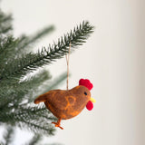This delightful handcrafted chicken felt ornament is suspended on a lush green Christmas tree branch, embodying a warm, joyful atmosphere. The brown chicken ornament features touches of orange, a bright red comb, and a small yellow beak, held by a rustic jute string. It's a unique Christmas tree ornament.