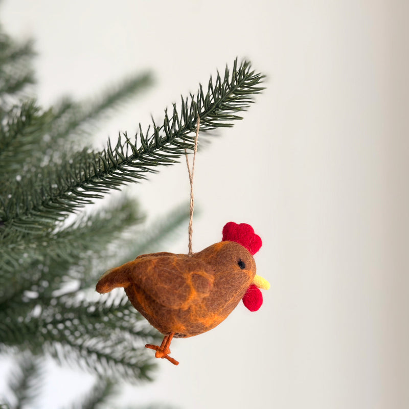This delightful handcrafted chicken felt ornament is suspended on a lush green Christmas tree branch, embodying a warm, joyful atmosphere. The brown chicken ornament features touches of orange, a bright red comb, and a small yellow beak, held by a rustic jute string. It's a unique Christmas tree ornament.