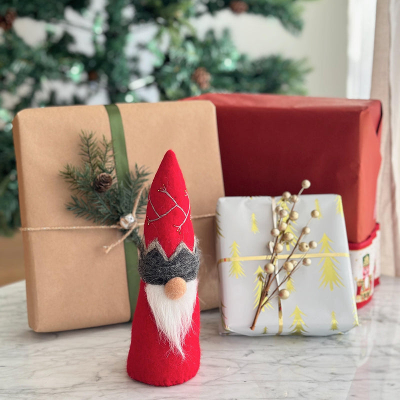 A charming red wool felt Christmas gnome ornament stands prominently in the foreground, creating a cozy, festive scene with wrapped gifts and a blurred Christmas tree in the background. This handcrafted felt gnome features a vibrant red hat with decorative stitching, a gray brim, and a soft white beard. Various wrapped gifts, including tan, white, and red boxes, complete the warm holiday setting.