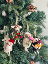 Three unique felt sheep Christmas ornaments are suspended from the branches of a green Christmas tree adorned with pine cones, creating a joyful and festive atmosphere. Each handcrafted felt sheep ornament is suspended by a jute string. One brown sheep wears a green wreath with red berries, a white sheep dons a red Santa hat, and a cream sheep holds a bouquet of pink, yellow, and green flowers.