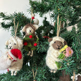 Three adorable handmade felt sheep Christmas ornaments are charmingly displayed on lush green Christmas tree branches. Each unique felt sheep ornament, suspended by a rustic jute string, features distinctive details. One white sheep wears a festive red Santa hat, another brown-faced sheep is adorned with green foliage and red berries, and the third brown-faced sheep holds a bouquet of pink and yellow flowers.