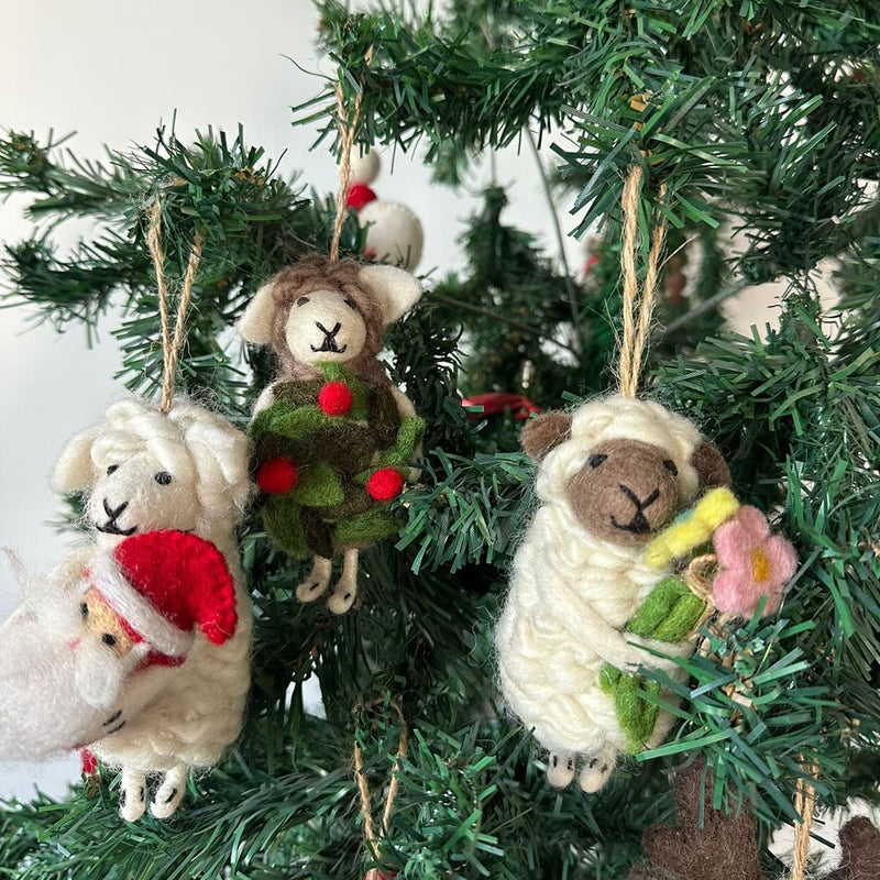 Three adorable handmade felt sheep Christmas ornaments are charmingly displayed on lush green Christmas tree branches. Each unique felt sheep ornament, suspended by a rustic jute string, features distinctive details. One white sheep wears a festive red Santa hat, another brown-faced sheep is adorned with green foliage and red berries, and the third brown-faced sheep holds a bouquet of pink and yellow flowers.