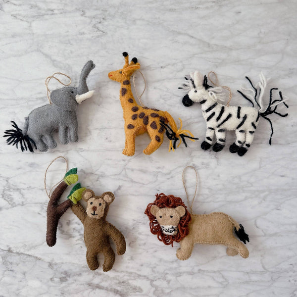 A playful set of five felt Christmas safari animal ornaments rests on a marble background. The collection includes a gray elephant felt ornament with white tusks, a yellow giraffe felt ornament with brown spots, and a black and white striped zebra felt ornament. A brown monkey felt ornament hangs from a branch with green leaves, while a tan lion felt ornament displays a brown yarn mane. Each handcrafted felt ornament includes a jute string for hanging.