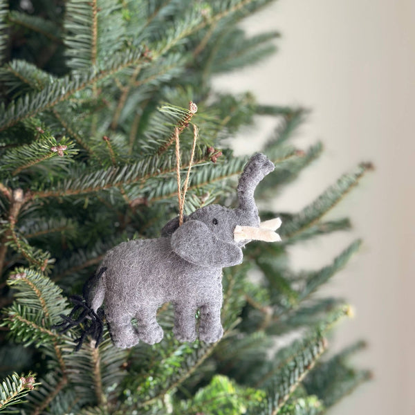 A felt elephant Christmas ornament is nestled among the vibrant green branches of a Christmas tree. This charming wool felt elephant ornament is crafted from soft gray felt, featuring a curled trunk, small tusks, floppy ears, and a playful black fringe tail. A jute string is attached at the top, ready to hang and add a touch of wild charm to holiday decor.