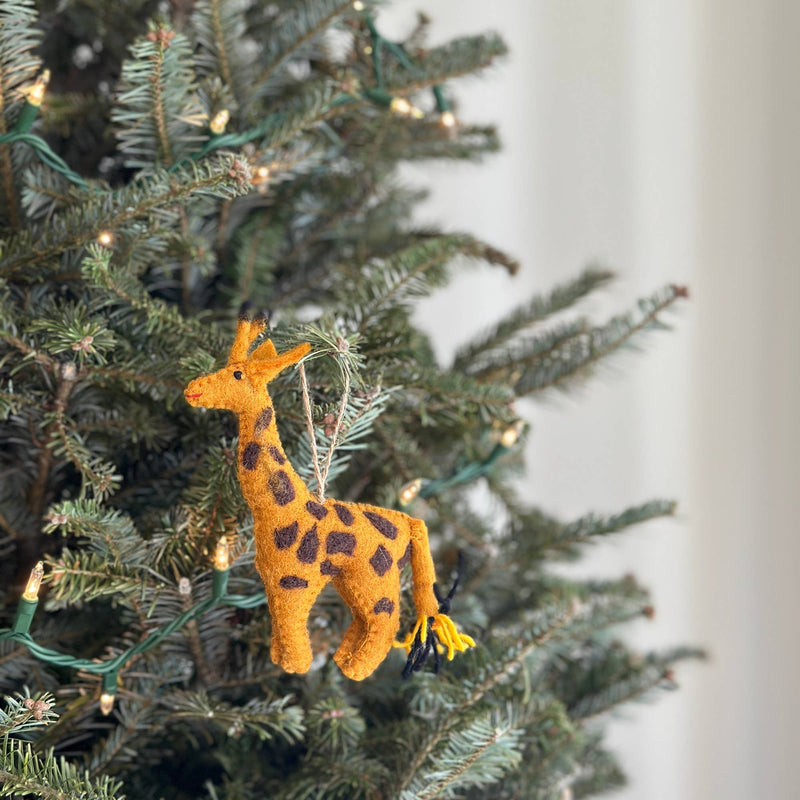 A single felt Christmas giraffe ornament hangs on a green Christmas tree adorned with small white lights. The handmade felt giraffe ornament is light orange with brown spots, complete with a jute string for hanging, creating a warm, inviting safari theme.