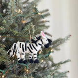A white wool felt zebra ornament, part of a set of safari animal felt ornaments, hangs festively from a Christmas tree adorned with soft, glowing lights. This unique Christmas ornament features distinctive black stripes, a black wool felt mane and legs, and a white string tail. Its ears have pink wool felt insides, with a jute string for hanging that blends seamlessly with the green branches.