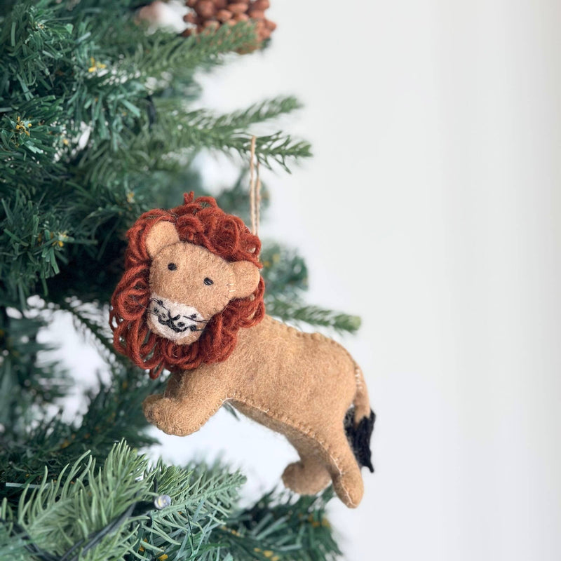 A felt lion Christmas ornament, part of a safari animal set, hangs from a green Christmas tree branch with pine cones visible. The charming lion features a tan felt body with visible stitching, a striking reddish-brown yarn mane, small black eyes, a white muzzle, and stitched whiskers, giving it a friendly appearance. A black yarn tail extends from its back, adding to its unique holiday charm.