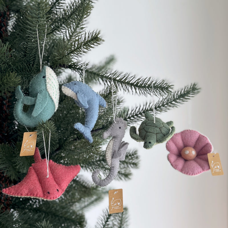 A set of six unique felt ocean creature Christmas ornaments hangs from a festive Christmas tree against a soft, neutral backdrop. The handmade wool felt ornaments include a light blue shark, a light blue streamlined dolphin, a gray textured seahorse, a dark green patterned turtle, a red flat stingray, and a pink pearl. White cotton strings secure each ornament, which also features a small cardboard tag.