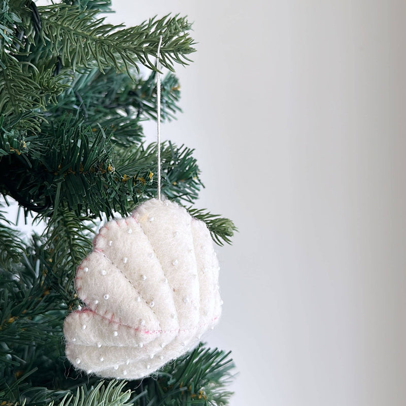 A white wool felt seashell ornament from the Ocean Creatures Felt Christmas Ornament set hangs on a green Christmas tree. This soft, spiral-shaped ornament features subtle pink stitching along its edges and small white beads detailing its ridges, with a white cotton string for hanging.