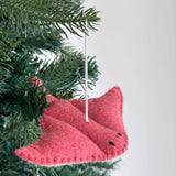 A red wool felt stingray Christmas ornament, part of an ocean creatures set, hangs from a green Christmas tree branch against a white background. This handmade felt ornament features a textured surface, delicate black eyes, and is suspended by a white cotton string, adding a festive touch.