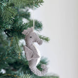 A gray and white wool felt seahorse Christmas ornament hangs gracefully from a green Christmas tree branch. This unique ocean creature ornament, part of the Felt Christmas Ornament Set, features a classic seahorse shape with a curved tail, snout, and fins. Its soft, endearing wool felt texture adds a touch of handmade holiday warmth, suspended by a white cotton string.
