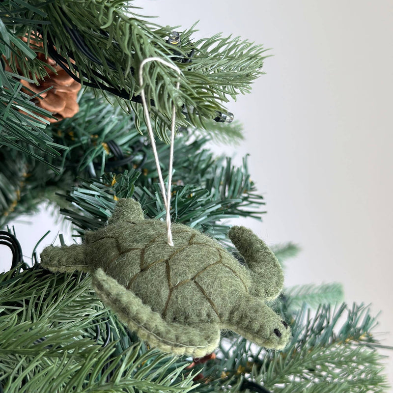 A muted green wool felt turtle Christmas ornament from the Ocean Creatures set hangs on a festive pine Christmas tree branch. The turtle features a segmented shell with detailed stitching and small, protruding flippers, suspended by a white cotton string.