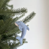 A light blue wool felt dolphin ornament from the Ocean Creatures Felt Christmas Ornament Set hangs on a green Christmas tree branch against a soft white background. This unique Christmas ornament features a smooth, rounded body with a lighter peach front, small black eyes, and a thin white cotton string. Its handcrafted appearance and slightly fuzzy texture add an endearing charm.