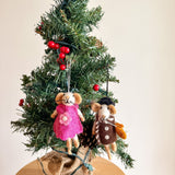 A set of two needle felted mouse Christmas ornaments, depicting a 'Little Sister' and 'Little Brother' mouse, hang on a small Christmas tree. The tree, decorated with red berries and mini lights, sits on a burlap-wrapped wooden base. The light-beige sister mouse wears a pink dress with a flower, while the brother mouse wears a dark brown coat, scarf, and black hat, holding a carrot. These 3D organic wool felt ornaments feature pointed ears and thread whiskers.