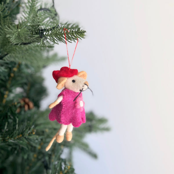 A charming handmade felt Little Sister mouse Christmas ornament hangs gracefully from a green Christmas tree. This delightful needle felted mouse, with a cream-colored body, wears a vibrant pink dress and a jaunty red hat. A red cotton string is attached, ready for hanging on the festive boughs.