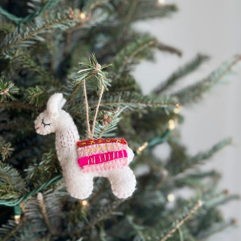 A handcrafted felt llama Christmas ornament hangs on a warmly lit Christmas tree. This charming white llama ornament features a colorful saddle in pink, brown, and yellow, with delicate stitched eyes and a red mouth. It is suspended by a jute string, blending into the festive, blurred background.