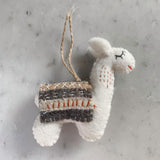 A cream-colored felt llama Christmas ornament is presented on a mottled gray surface. This charming felted ornament features closed black eyes and a small red stitched smile. It wears a saddle blanket detailed with striped brown, beige, and gray patterns, each adorned with unique stitches. A jute string is attached for hanging, completing its handcrafted look.