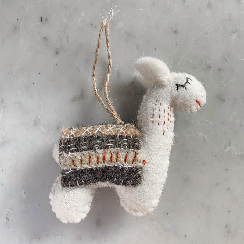 A cream-colored felt llama Christmas ornament is presented on a mottled gray surface. This charming felted ornament features closed black eyes and a small red stitched smile. It wears a saddle blanket detailed with striped brown, beige, and gray patterns, each adorned with unique stitches. A jute string is attached for hanging, completing its handcrafted look.