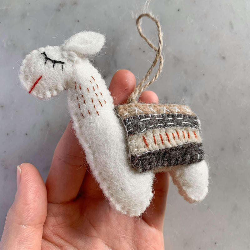 A female hand gently holds one white felt llama Christmas ornament against a mottled gray and white surface. This handcrafted felt ornament features a jute string loop for hanging. The white llama has a rectangular saddle with brown, gray, and white stripes accented with red. Its face shows closed black eyes and a red stitched mouth, with small red stitches highlighting its body.