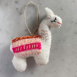 A charming white felt llama ornament rests on a mottled gray and white surface. This handmade felt llama ornament features a detailed white felt body, accented with a decorative brown and pink saddle, stitched eyes, and a red stitched mouth. Upright ears and a jute string at the top complete the design, ready for hanging.