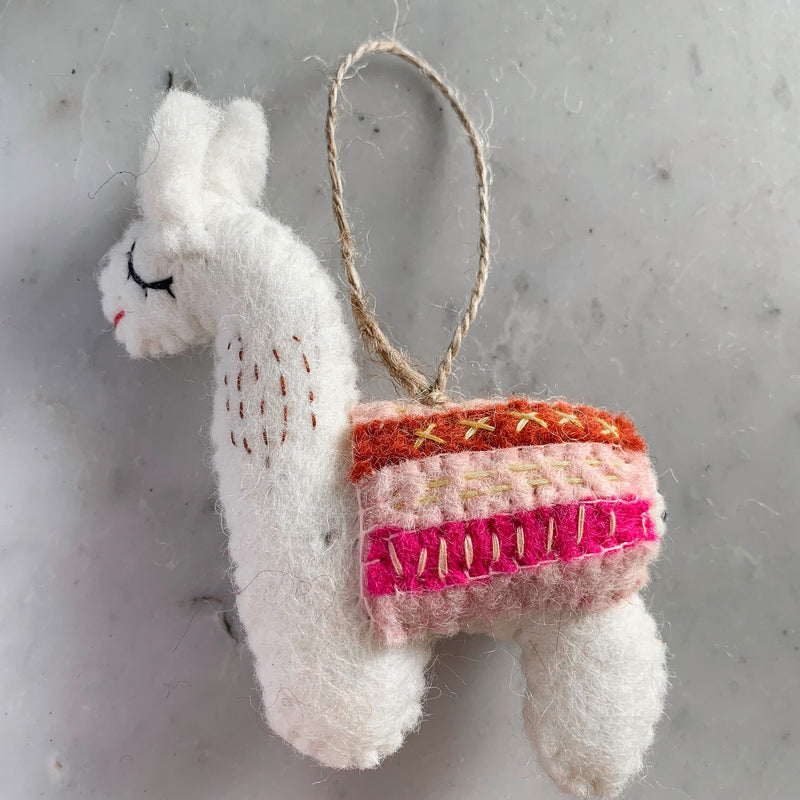 A white felt Christmas llama ornament is displayed against a mottled gray and white background. The handmade felt llama features black stitched eyes and a small red mouth. Its neck has dark brown stitching, and a multi-colored saddle pad with pink, orange, and light brown striped details adorns its back. A jute string extends from the top for hanging.